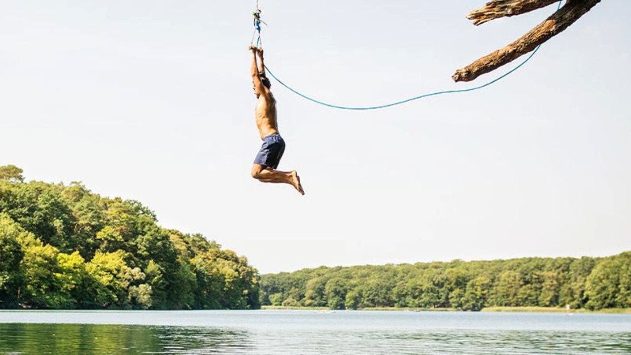 Imagem de uma paisagem de natureza, com um lago azul e várias árvores verdes em seu redor. Do lado direito da imagem podemos ver um homem pendurado numa corda amarrada a uma árvore, que se prepara para se atirar ao lago. O homem veste apenas uns calções de banho.