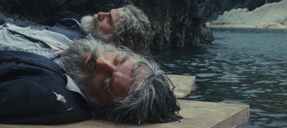 Um homem com barba grisalha, a usar um fato rasgado, está deitado de lado, aparentemente inconsciente ou morto, sobre uma plataforma de madeira à beira de um lago ou rio, com rochas e vegetação ao fundo. A expressão do homem é serena e há outra figura parcialmente visível e desfocada atrás dele. Um homem com barba grisalha, a usar um fato rasgado, está deitado de lado, aparentemente inconsciente ou morto, sobre uma plataforma de madeira à beira de um lago ou rio, com rochas e vegetação ao fundo. A expressão do homem é serena e há outra figura parcialmente visível e desfocada atrás dele.