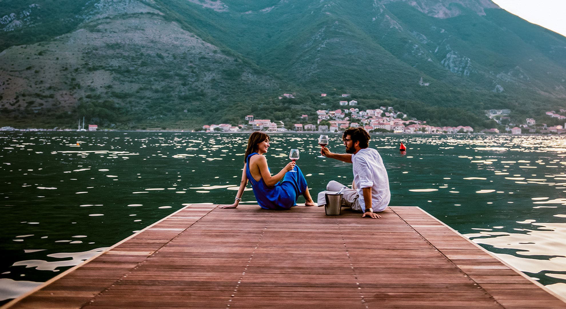 Homem e mulher a brindar num deck de madeira sobre um lago, com um monte verde com casas como pano de fundo.