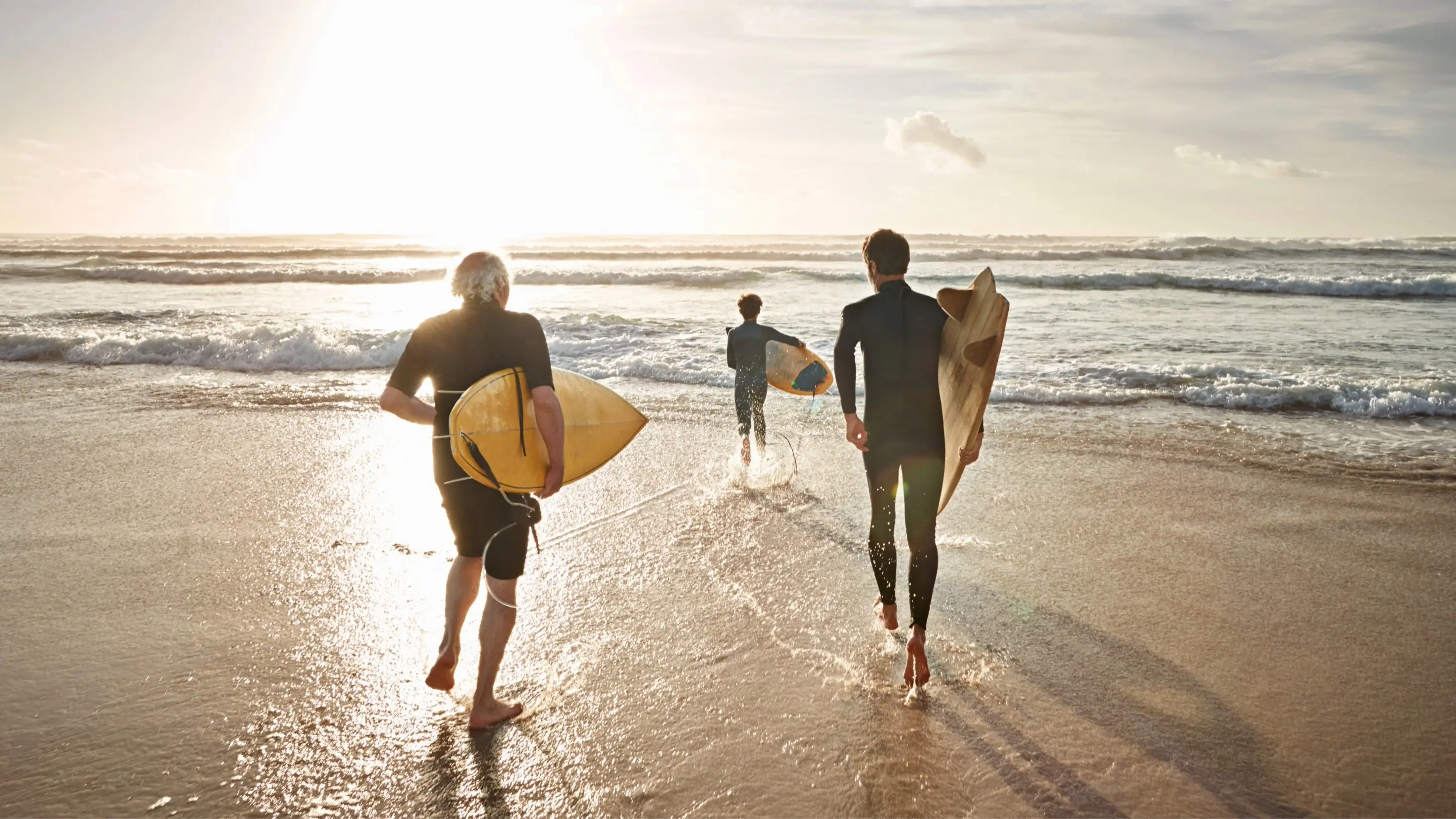 Três pessoas, cada uma delas com a sua prancha e fato de surf, prontaspara entrar no mar numa praia. Do lado esquerdo pode observar-se o reflexo do sol no mar e na areia.