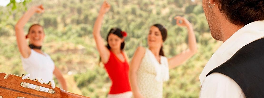 Trois femmes dansent en plein air les bras levés, tandis qu’un musicien observe au premier plan, dans un décor naturel.