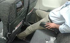 Photo of a man sitting by the window of an airplane, on a two-seat row, with his left leg stretched out under the front seat.