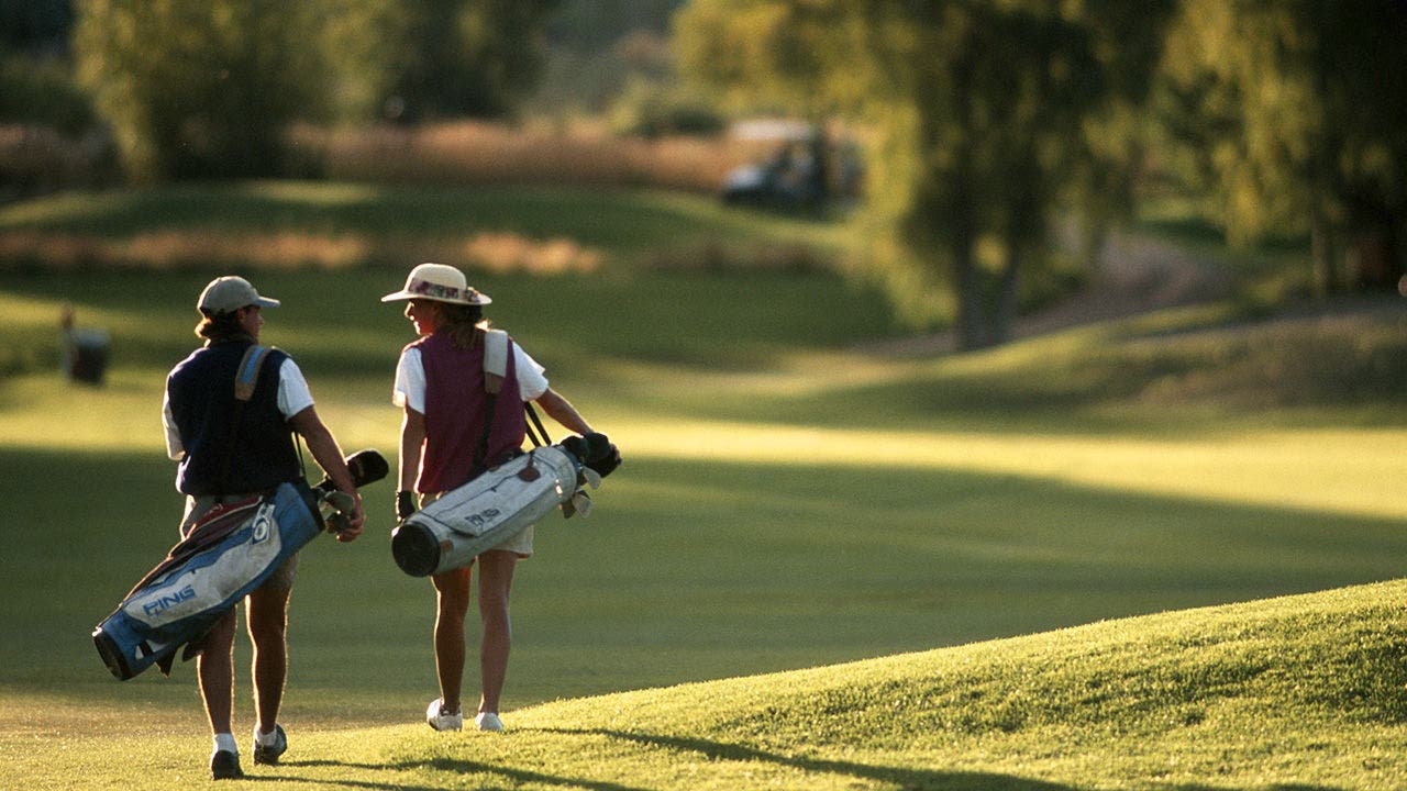 Two people walking across a golf course carrying golf bags with equipment.