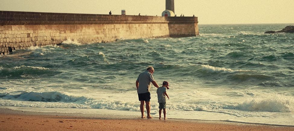 Idoso caminha junto à praia com uma criança, ambos de mãos dadas e próximos da água do mar; ao fundo, vê-se um paredão e um farol junto ao oceano. Idoso caminha junto à praia com uma criança, ambos de mãos dadas e próximos da água do mar; ao fundo, vê-se um paredão e um farol junto ao oceano.