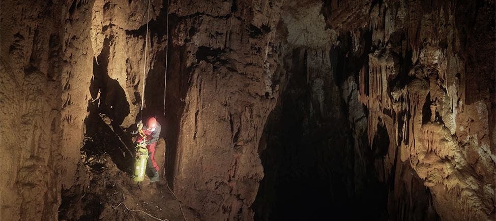 Espeleólogo com capacete e roupa protetora ilumina uma zona de uma gruta extensa, suspenso por cordas entre as paredes rochosas, numa área com pouca luz natural. Espeleólogo com capacete e roupa protetora ilumina uma zona de uma gruta extensa, suspenso por cordas entre as paredes rochosas, numa área com pouca luz natural.