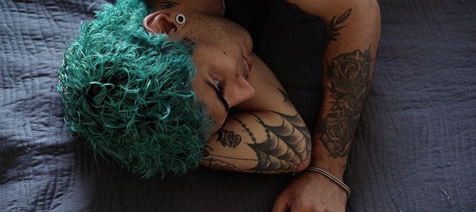 Jovem de cabelo pintado de verde deitado de lado sobre uma colcha escura, que parece ser de uma cama, com vários piercings e tatuagens visíveis no braço e ombro, numa pose relaxada. Jovem de cabelo pintado de verde deitado de lado sobre uma colcha escura, que parece ser de uma cama, com vários piercings e tatuagens visíveis no braço e ombro, numa pose relaxada.