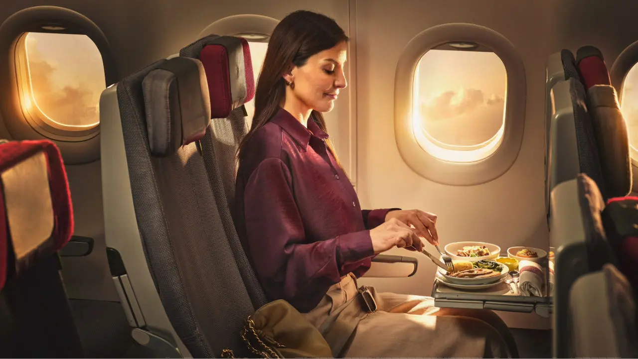 Une femme assise près du hublot d’un avion prend un repas pendant le vol, éclairée par la lumière du soleil, avec le siège à côté d’elle vide.