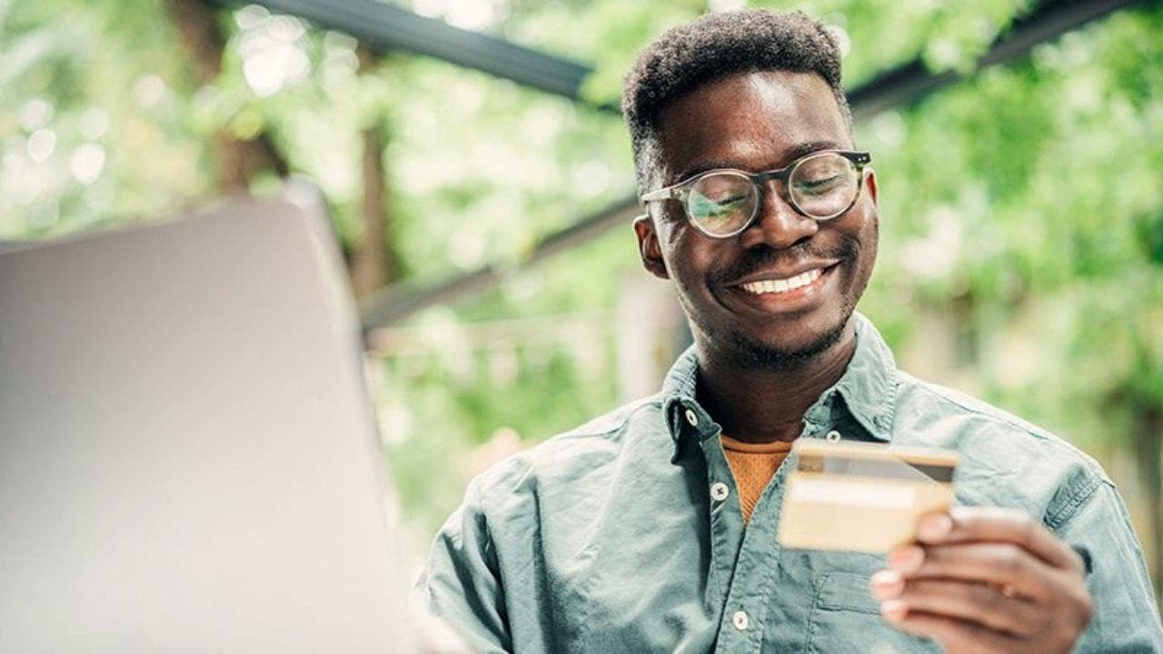 A imagem mostra um homem ao computador e a sorrir para um cartão de crédito que tem na sua mão esquerda. Por detrás dele, avista-se uma área verde, de arbustos. A imagem mostra um homem ao computador e a sorrir para um cartão de crédito que tem na sua mão esquerda. Por detrás dele, avista-se uma área verde, de arbustos.
