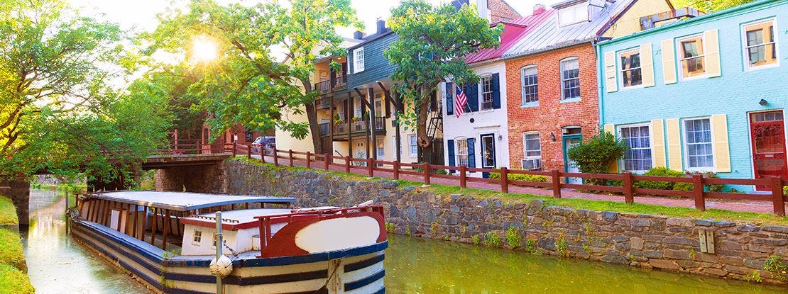 Casas históricas coloridas à beira de um canal arborizado, com um barco atracado na água e luz do sol entre as árvores.