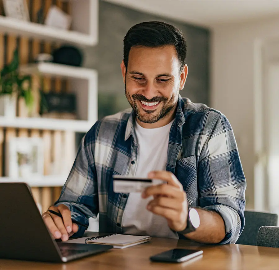 Um homem moreno jovem, sorri para um cartão de crédito. À sua frente encontra-se um caderno de apontamentos e um computador portátil.