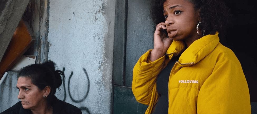 Duas mulheres, uma com casaco amarelo ao telemóvel e expressão apreensiva, outra sentada ao fundo com ar sério, à porta de um edifício degradado, paredes grafitadas e ambiente urbano. Duas mulheres, uma com casaco amarelo ao telemóvel e expressão apreensiva, outra sentada ao fundo com ar sério, à porta de um edifício degradado, paredes grafitadas e ambiente urbano.