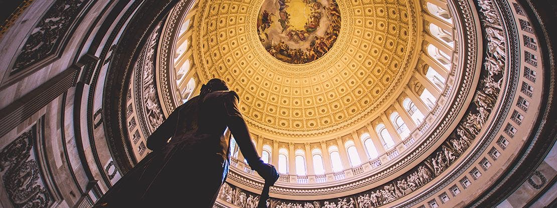 Vue intérieure de la coupole du Capitole des États-Unis, prise vers le haut, avec une statue au premier plan et des ornements dorés.