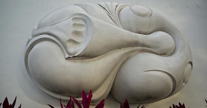 Escultura abstrata em relevo de cor branca com formas orgânicas e curvas, fixada numa parede clara, com folhas vermelhas surgindo na base da imagem.