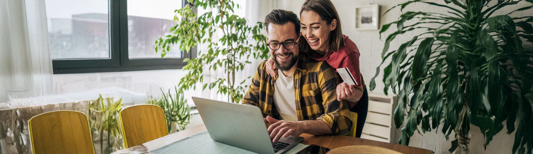 Um homem sentado a uma secretária a utilizar um computador portátil, com uma mulher atrás dele a apoiar-se no seu ombro e a segurar um cartão bancário.