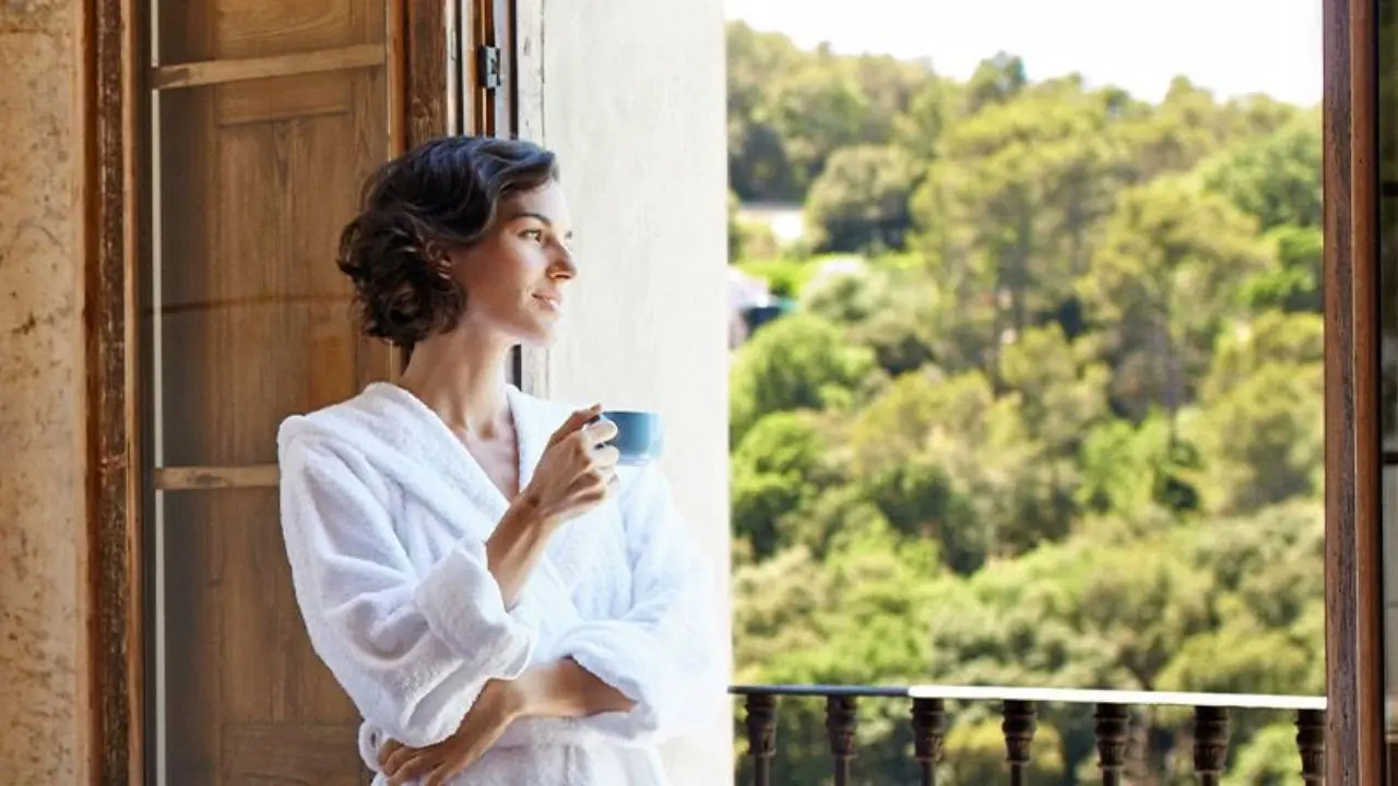 A imagem mostra uma senhora na varanda a apreciar a vista. Lá fora, avistam-se árvores. A senhora veste um robe branco e tem uma chávena de café azul na mão direita.