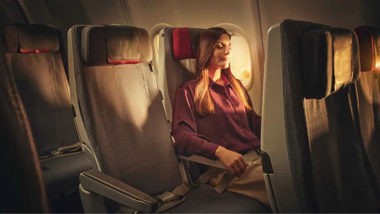 A woman seated by an airplane window relaxes during the flight, illuminated by sunlight entering the cabin, with the seat next to her empty.