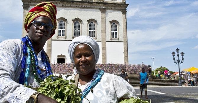 Pessoas em trajes tradicionais brancos com colares coloridos segurando ramos verdes em frente a uma igreja histórica em Salvador, Bahia, Brasil.