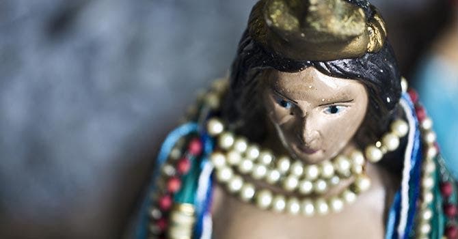 Close-up de uma pequena escultura com colares de pérolas e fios coloridos ao redor do pescoço, em foco, contra um fundo desfocado.