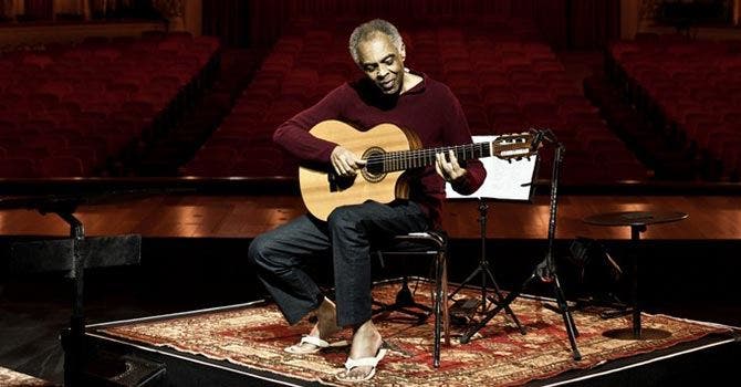 Pessoa descalça sentada numa cadeira tocando guitarra em cima de um tapete, sozinha num palco iluminado diante de uma plateia vazia.