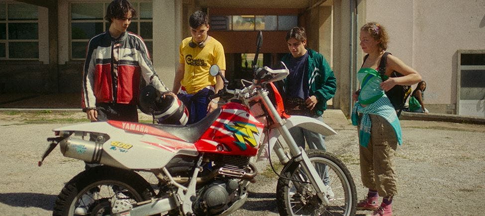 Quatro jovens em pé em redor de uma mota Yamaha estacionada ao ar livre, conversando e sorrindo; arquitetura escolar visível ao fundo. Quatro jovens em pé em redor de uma mota Yamaha estacionada ao ar livre, conversando e sorrindo; arquitetura escolar visível ao fundo.