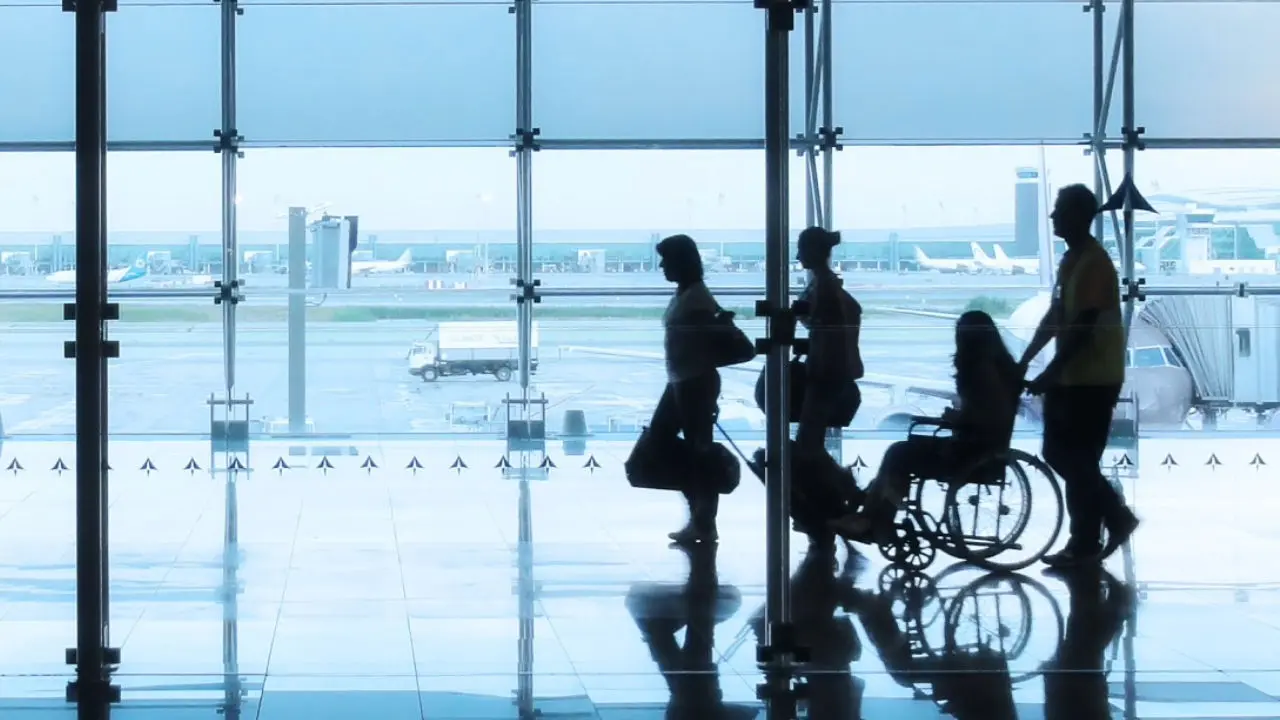 Imagem em contraluz de quatro pessoas a deslocarem-se num aeroporto. Duas delas encontram-se a carregar a sua bagagem, outra encontra-se numa cadeira de rodas que está a ser guiada pela última pessoa.