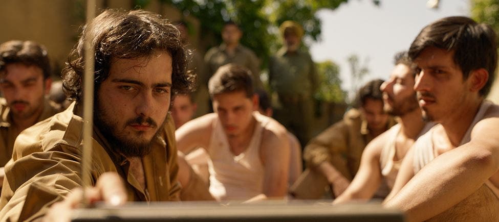 Grupo de jovens sentados num pátio ao ar livre, com expressões sérias, alguns de camisola interior, outros de farda castanha; dois homens de uniforme observam de pé ao fundo. Grupo de jovens sentados num pátio ao ar livre, com expressões sérias, alguns de camisola interior, outros de farda castanha; dois homens de uniforme observam de pé ao fundo.