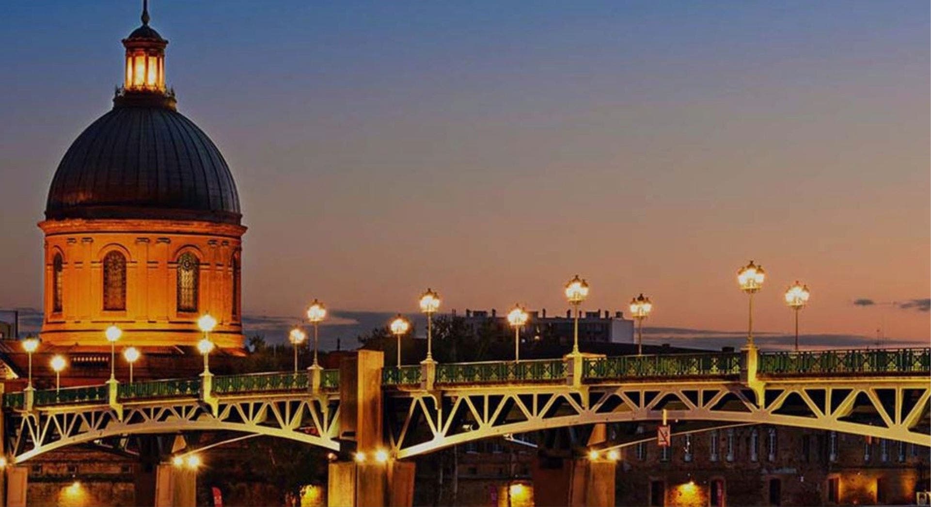 Cúpula iluminada e ponte metálica com candeeiros acesos ao entardecer em Toulouse, França, sob um céu degradê azul e laranja.