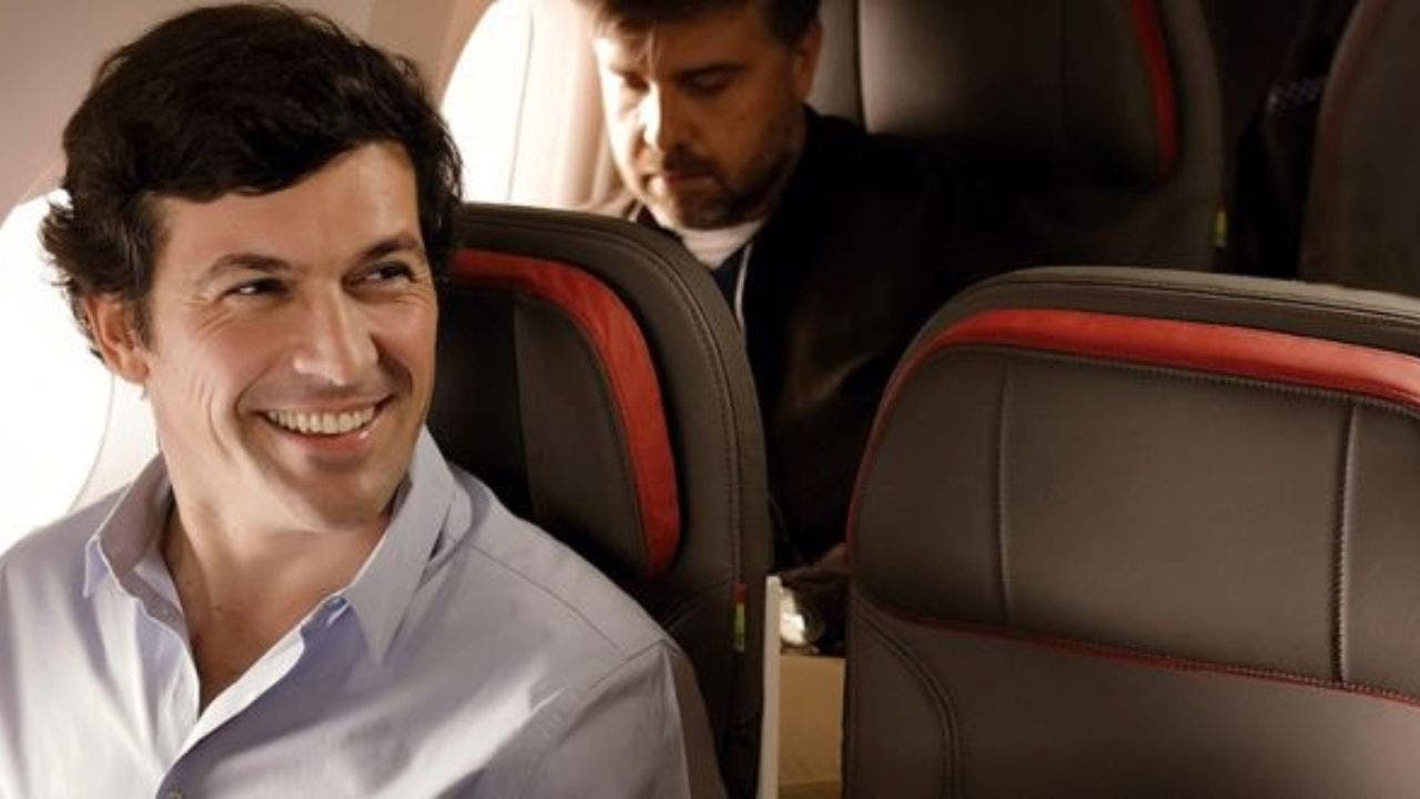Plano aproximado de um senhor, sentado à janela de um avião. É moreno e veste uma camisa azul-clara, está a sorrir para o seu lado esquerdo.