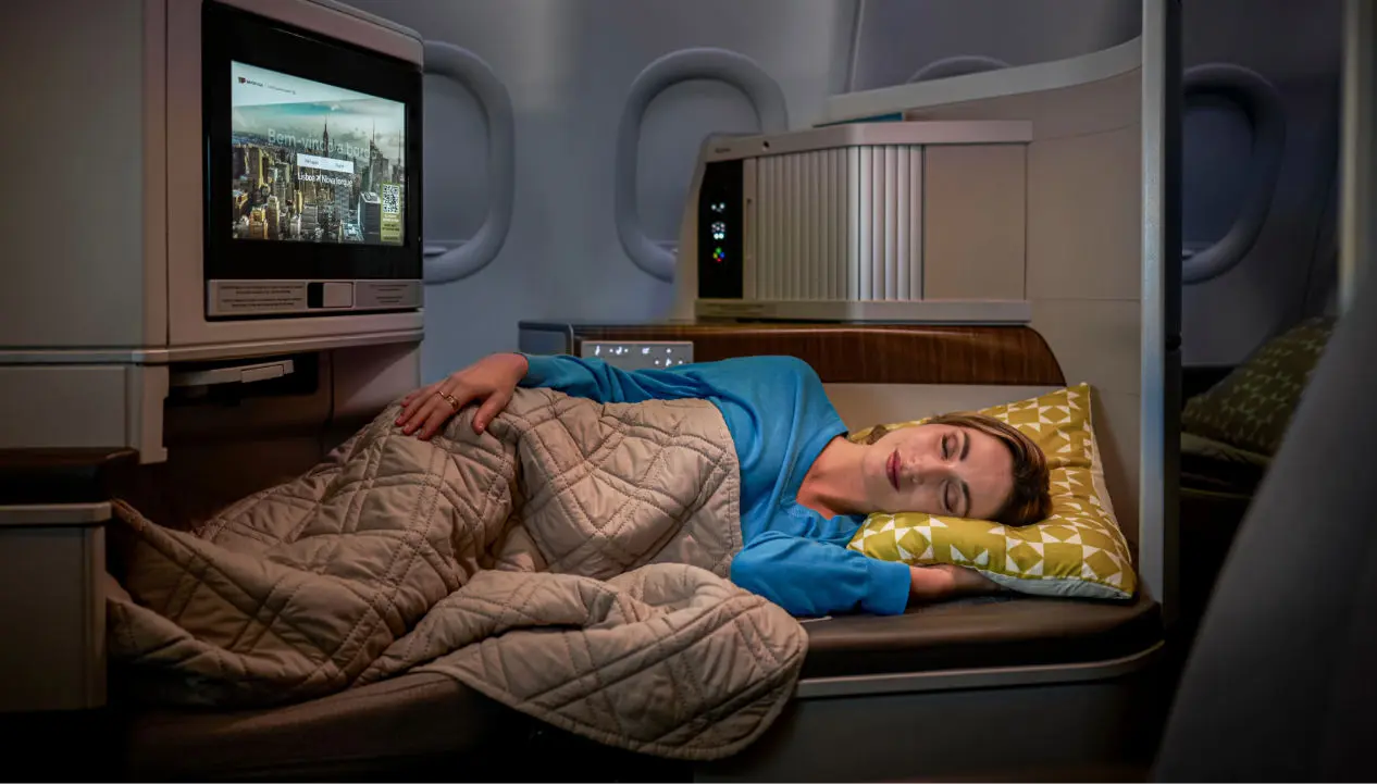 Une femme dort confortablement allongée dans une cabine d’avion, couverte d’une couette, sur un siège qui se transforme en lit, avec un écran de divertissement à côté. Une femme dort confortablement allongée dans une cabine d’avion, couverte d’une couette, sur un siège qui se transforme en lit, avec un écran de divertissement à côté.