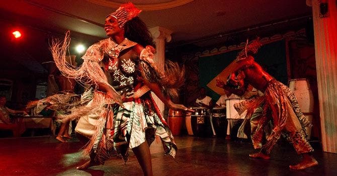 Dançarinos em trajes tradicionais afro-brasileiros com fibras e estampas coloridas realizando uma apresentação enérgica num palco iluminado em tons vermelhos, acompanhados por tambores ao fundo.