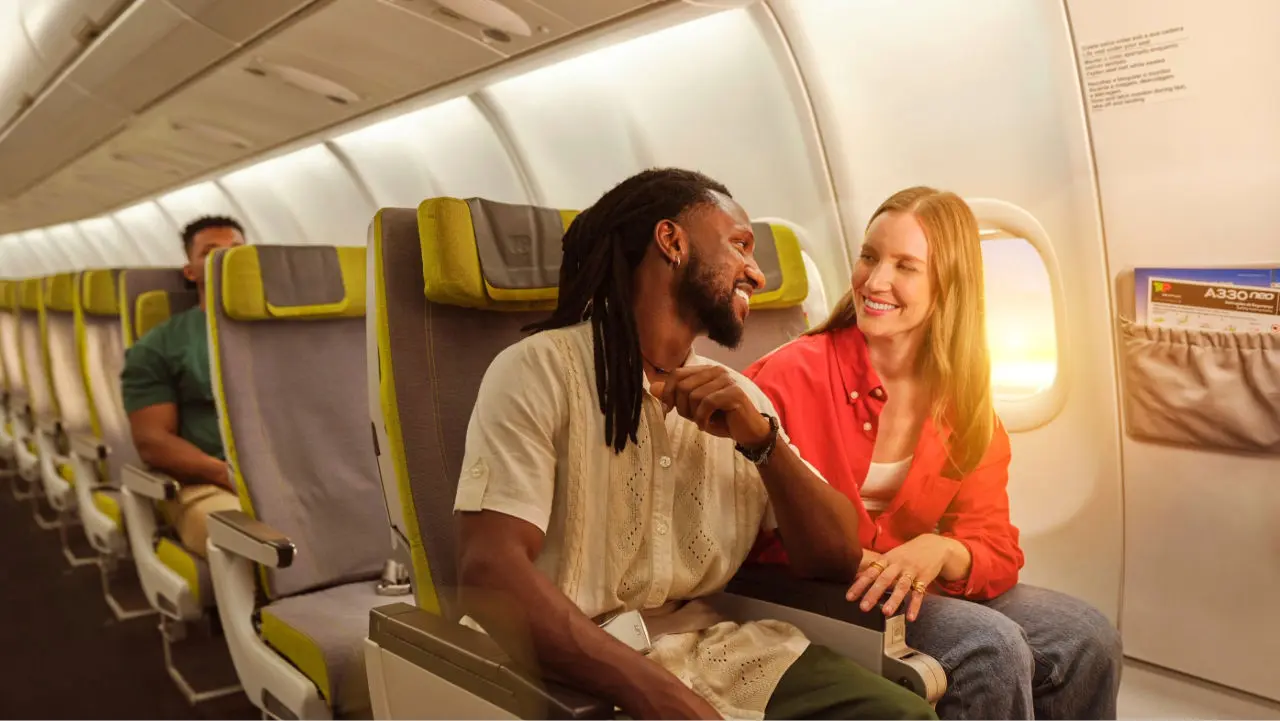 Un couple assis côte à côte dans un avion discute et sourit pendant le vol, avec d’autres sièges autour apparemment libres.