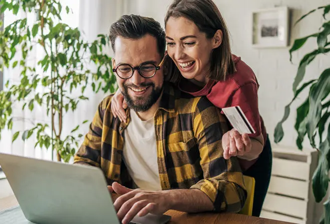 Um homem sentado a uma secretária a utilizar um computador portátil, com uma mulher atrás dele a apoiar-se no seu ombro e a segurar um cartão bancário.